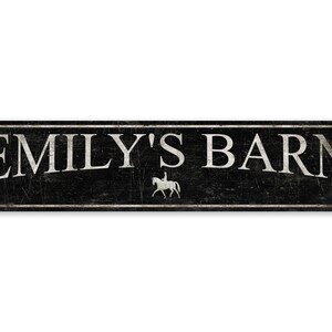 May include: Black metal sign with white text that reads "Emily's Barn". A white silhouette of a person riding a horse is centered below the text.