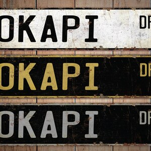 May include: Three vintage-style metal signs with the word "OKAPI" and "DR" in bold, distressed lettering. The signs are in black, white, and yellow, with a rustic, weathered appearance, suggesting a street sign theme.