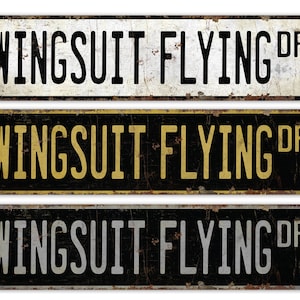 May include: Three vintage-style metal signs with the text "WINGSUIT FLYING DR" in black lettering on a white, gold, and silver background. The signs have a distressed, rusty look.