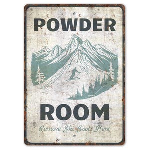 以下が含まれることがあります： 長方形の金属製の看板で、使い古したような仕上がりです。「POWDER ROOM」の文字が上下にあり、スキーヤーがいる山の風景が描かれています。「Remove Ski Boots Here」の文字が下にあります。