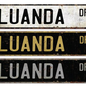 May include: Three vintage-style metal street signs with the text "LUANDA DR" in black lettering on a white, yellow, and gray background. The signs are weathered and have a distressed look.