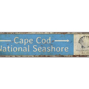 May include: A rectangular metal sign with a distressed finish. The sign features the text "Cape Cod National Seashore" in white, with an arrow pointing right. A seashell graphic and the text "Massachusetts Est. 1961" are on the right.