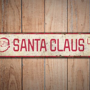May include: A vintage-style metal sign with a red border and white background. The sign reads "SANTA CLAUS LN" in red letters with a cartoon image of Santa Claus in the upper left corner.