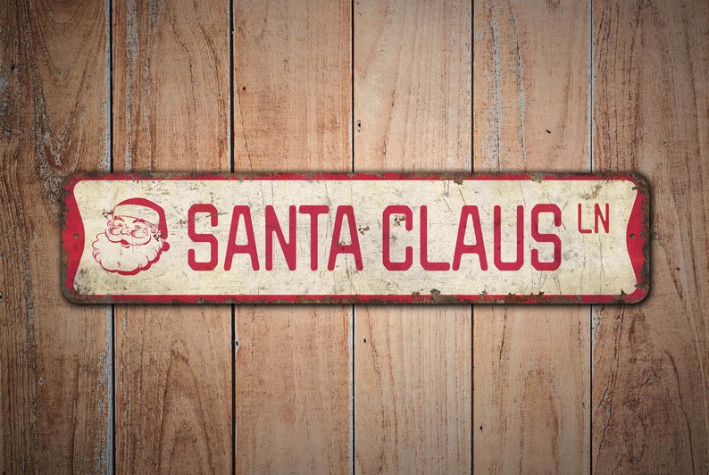 May include: A vintage-style metal sign with a red border and white background. The sign reads "SANTA CLAUS LN" in red letters with a cartoon image of Santa Claus in the upper left corner.
