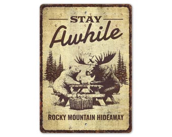 May include: Aged metal sign with a sepia tone. It depicts a bear and a moose at a picnic table, with the text "Stay Awhile" and "Rocky Mountain Hideaway". The sign has a distressed finish, giving it a vintage appearance.