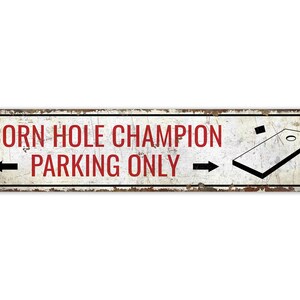 May include: Rectangular metal sign with a distressed finish. The sign reads "CORN HOLE CHAMPION PARKING ONLY" in red letters. A black arrow points left, and a cornhole board graphic is on the right.
