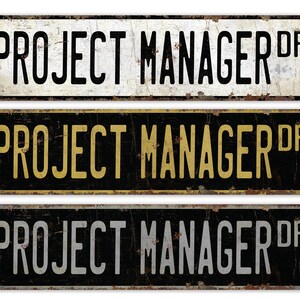May include: Three vintage-style metal signs with the text "PROJECT MANAGER DR" in different colors. The top sign has white text on a white background, the middle sign has yellow text on a black background, and the bottom sign has gray text on a black background.