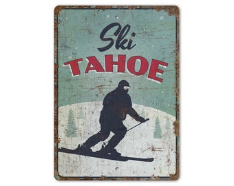 May include: A vintage-style metal sign with a distressed blue background and red text that reads "Ski Tahoe". A silhouette of a skier is shown in the center of the sign, skiing down a snowy slope.