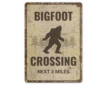 May include: A vintage-style metal sign with a distressed, beige background. The sign features a silhouette of Bigfoot walking, with the text "BIGFOOT CROSSING NEXT 3 MILES".