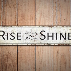 以下が含まれることがあります： 木製の背景に、「Rise and Shine」と書かれた黒い文字の白い金属製の看板