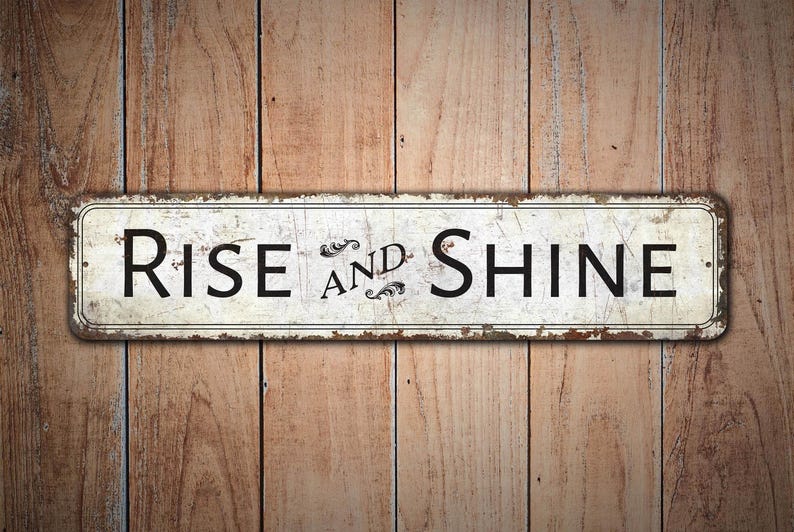 以下が含まれることがあります： 木製の背景に、「Rise and Shine」と書かれた黒い文字の白い金属製の看板