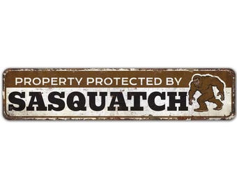 May include: A brown metal sign with white lettering that reads "PROPERTY PROTECTED BY SASQUATCH". A cartoon image of a brown sasquatch is on the right side of the sign.