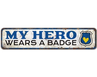 May include: A white metal sign with blue trim and the text "MY HERO WEARS A BADGE" in black. A blue and yellow shield with a yellow heart is on the right side of the sign.
