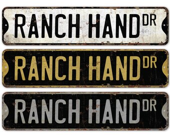 May include: Three vintage-style metal signs with the text "RANCH HAND DR" in black lettering on a white, gold, and silver background. The signs have a distressed, rusty look.