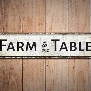 Puede incluir: Un letrero de metal blanco con el texto "FARM to TABLE" en letras negras y un peque&ntilde;o dise&ntilde;o de hojas entre las palabras.