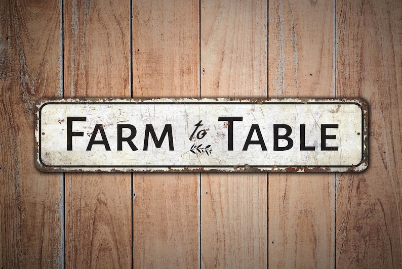 Puede incluir: Un letrero de metal blanco con el texto "FARM to TABLE" en letras negras y un peque&ntilde;o dise&ntilde;o de hojas entre las palabras.