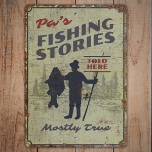 以下が含まれることがあります： ヴィンテージ風の金属製サイン。赤と黒の文字で「Pa's FISHING STORIES」と書かれ、大きな魚を持った漁師のイラストが描かれています。「TOLD HERE」と「Mostly True」の文字も含まれています。レトロな外観です。