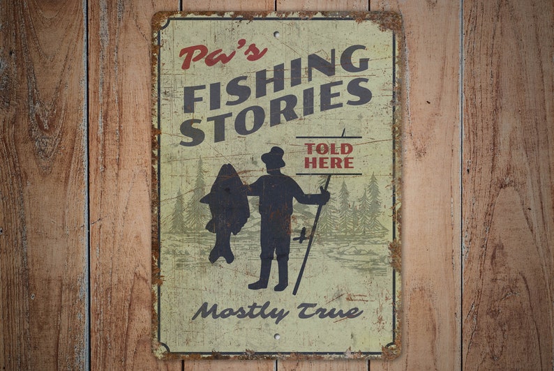 以下が含まれることがあります： ヴィンテージ風の金属製サイン。赤と黒の文字で「Pa's FISHING STORIES」と書かれ、大きな魚を持った漁師のイラストが描かれています。「TOLD HERE」と「Mostly True」の文字も含まれています。レトロな外観です。