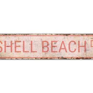 May include: A vintage-style metal sign with a white background and red text that reads "SHELL BEACH Dr". The sign has a distressed, rusty look.