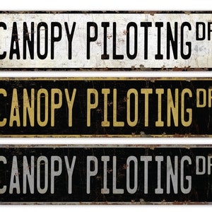 May include: Three vintage-style metal signs with the text "CANOPY PILOTING DR" in black, yellow, and gray lettering on a black, white, and black background, respectively.