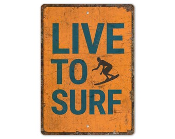 May include: Distressed orange metal sign with the words "LIVE TO SURF" in teal. A silhouette of a surfer on a wave is also in teal. The sign has a vintage appearance.