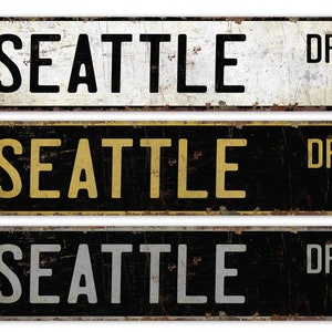 May include: Three vintage-style metal street signs with the text "SEATTLE DR" in black, gold, and silver lettering on a black background.