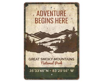 May include: A rectangular metal sign with a distressed finish. The sign features the text "ADVENTURE BEGINS HERE" in brown, above a silhouette of mountains and trees. Below, it reads "GREAT SMOKY MOUNTAINS National Park" and coordinates.