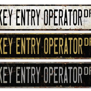 May include: Three vintage-style metal signs with the text "KEY ENTRY OPERATOR DR" in black, gold, and silver lettering on a black, white, and black background, respectively.