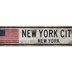 Op de afbeelding: Een vintage metalen bord met een verweerde witte achtergrond en zwarte tekst. Het bord heeft de tekst "NEW YORK CITY NEW YORK" en heeft een kleine Amerikaanse vlag in de linkerbovenhoek.