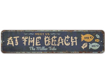 May include: A rectangular, distressed metal sign with the words "MEET US AT THE BEACH" in a stylized font. The sign features fish illustrations in yellow and blue, and the text "The Miller Tribe" is below.