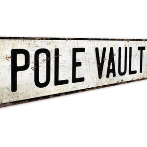 May include: A white metal sign with black text that reads "POLE VAULT DR". The sign has a distressed, vintage look.