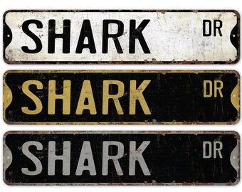 May include: Three vintage-style metal street signs with the text "SHARK DR" in black, gold, and silver lettering on a black background.