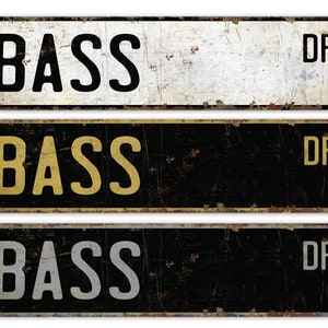 Puede incluir: Tres letreros de metal de estilo vintage con el texto "BASS DR" en diferentes colores. El letrero superior tiene texto negro sobre fondo blanco, el letrero del medio tiene texto amarillo sobre fondo negro y el letrero inferior tiene texto gris sobre fondo negro.