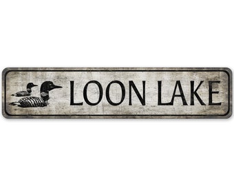 May include: A rustic, weathered sign with the text "LOON LAKE" in black lettering. The sign features a black silhouette of a loon with its chicks.