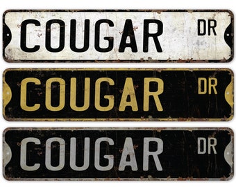May include: Three vintage-style metal street signs with the text "COUGAR DR" in black, gold, and silver lettering on a black background.