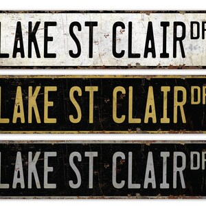 May include: Three vintage-style metal street signs with the text "LAKE ST CLAIR DR" in black, gold, and white lettering on a black, white, and black background, respectively.