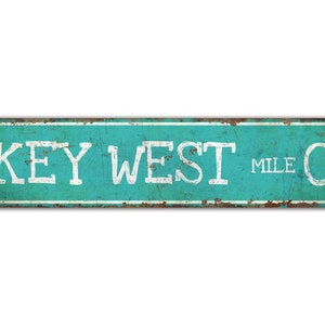 May include: A turquoise blue metal sign with white text that reads "KEY WEST MILE 0". The sign has a distressed, rusty look.