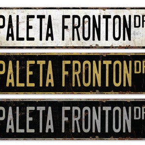May include: Three vintage-style metal street signs with the text "PALETA FRONTON DR" in black, gold, and white lettering on a black background.