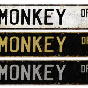 May include: Three vintage-style metal street signs with the text "MONKEY DR". The signs have a distressed look with rust and wear. The top sign is white with black text, the middle is black with yellow text, and the bottom is black with silver text.