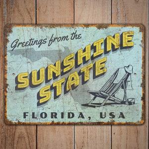 May include: Vintage metal sign with a distressed finish. The sign reads "Greetings from the SUNSHINE STATE FLORIDA, USA" in yellow and black lettering. A beach chair and hat are illustrated on the right side.