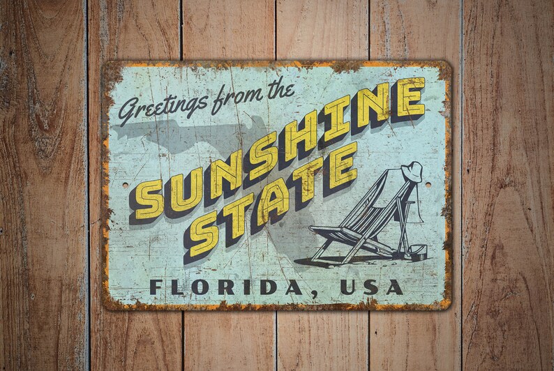 May include: Vintage metal sign with a distressed finish. The sign reads "Greetings from the SUNSHINE STATE FLORIDA, USA" in yellow and black lettering. A beach chair and hat are illustrated on the right side.
