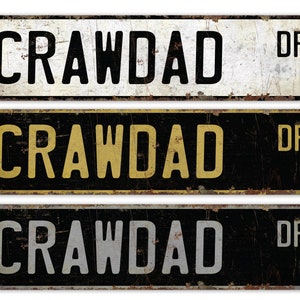 May include: Three vintage-style metal street signs with the text "Crawdad Dr." The top sign is white with black lettering, the middle sign is black with gold lettering, and the bottom sign is black with silver lettering.