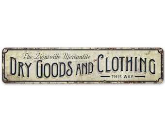 Op de afbeelding: Een vintage metalen bord met een witte achtergrond en zwarte tekst die "The Zionsville Mercantile Dry Goods and Clothing - This Way" luidt. Het bord heeft een verweerde, roestige uitstraling.