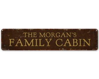 May include: A rectangular, distressed metal sign with the text "THE MORGAN'S FAMILY CABIN" in gold lettering. The sign has a brown background with a weathered appearance, suggesting an aged or vintage aesthetic. The sign has holes in each corner.