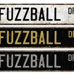 May include: Three vintage-style metal street signs with the text "Fuzzball Dr." The top sign has white background with black text, the middle sign has black background with gold text, and the bottom sign has black background with silver text.