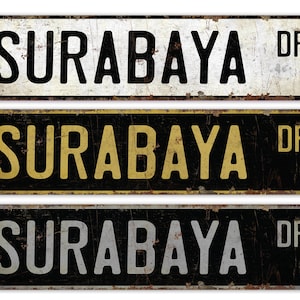 May include: Three vintage-style street signs with the text "SURABAYA DR". The top sign is white with black text, the middle sign is black with yellow text, and the bottom sign is black with silver text. All signs have a distressed, rusty appearance.
