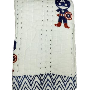 May include: White quilted blanket with a Captain America design. The blanket features a blue and red Captain America graphic and a blue zigzag pattern border. The blanket is stitched with black thread.