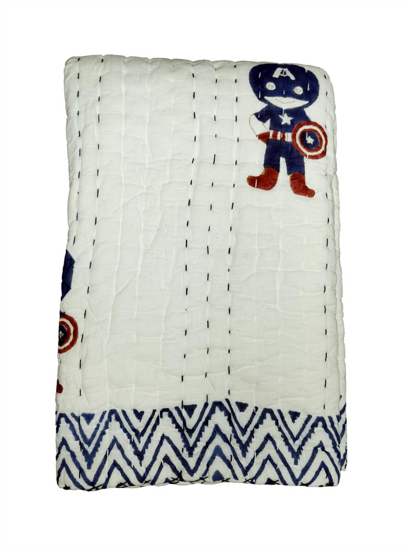 May include: White quilted blanket with a Captain America design. The blanket features a blue and red Captain America graphic and a blue zigzag pattern border. The blanket is stitched with black thread.