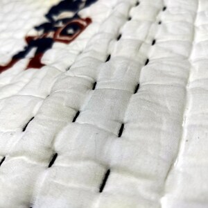 May include: Close-up of a white quilted fabric with black stitching. A partial image of a Captain America figure in red, white, and blue is visible in the upper left corner. The fabric has a textured, padded appearance.