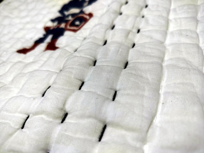 May include: Close-up of a white quilted fabric with black stitching. A partial image of a Captain America figure in red, white, and blue is visible in the upper left corner. The fabric has a textured, padded appearance.